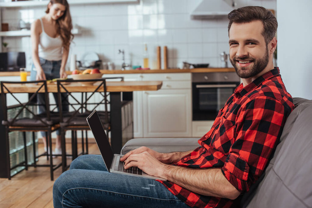 селективный фокус красивого фрилансера, улыбающегося в камеру во время работы над ноутбуком на кухне рядом с подругой на заднем плане
 - Фото, изображение