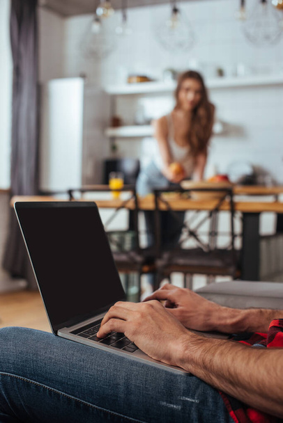 bijgesneden weergave van freelance typen op laptop met leeg scherm in de keuken in de buurt van vriendin op de achtergrond - Foto, afbeelding
