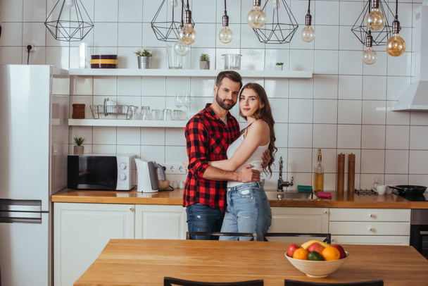 feliz jovem casal abraçando e olhando para a câmera enquanto em pé na cozinha moderna
 - Foto, Imagem