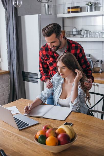 handsome man touching shoulders of attractive girlfriend looking at laptop and writing in notebook in kitchen - Фото, зображення