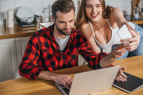 cheerful girl taking selfie with boyfriend sitting at kitchen table and working on laptop - Фото, зображення