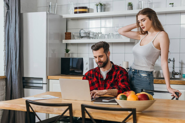 attractive girl standing near boyfriend sitting at kitchen table and working on laptop - Фото, изображение