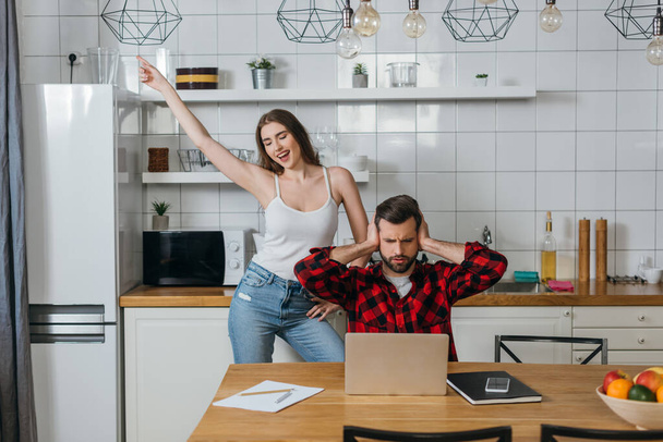 cheerful girl fooling around and dancings near boyfriend covering ears with hands while sitting near laptop and papers - Photo, Image