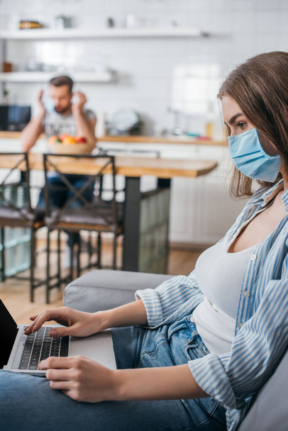 selective focus of young freelancer in medical mask working on laptop in kitchen near boyfriend on background - Fotografie, Obrázek