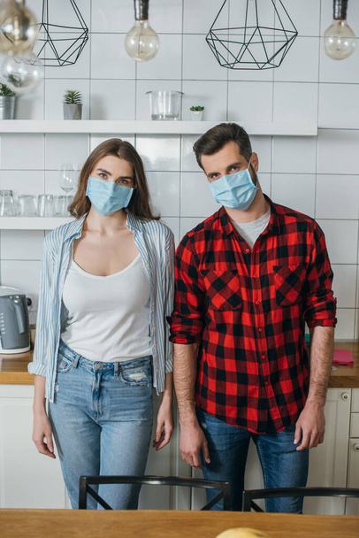 mladý pár v lékařských maskách při pohledu na kameru, zatímco stojí v moderní kuchyni - Fotografie, Obrázek