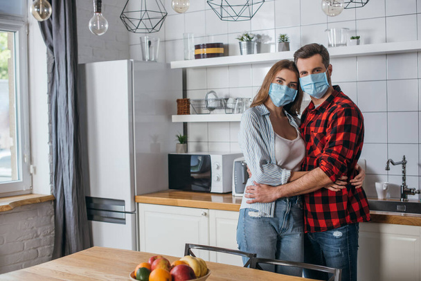 young couple in medical masks embracing and looking at camera in modern kitchen - Foto, Imagen