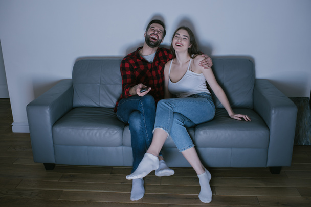 cheerful couple laughing while watching tv on sofa at home - Foto, Imagem