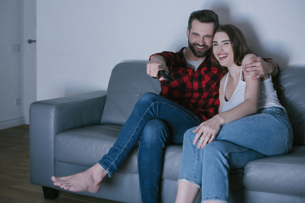 cheerful couple laughing and man pointing with finger while watching tv on sofa at home - Foto, afbeelding