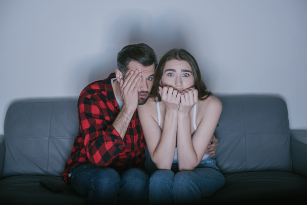 scared man covering eye with hand while watching movie near frightened girl - Фото, изображение