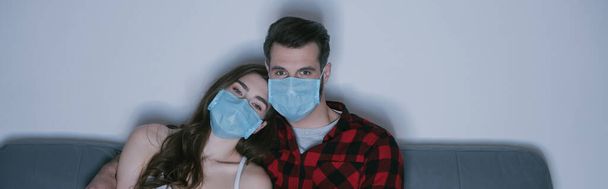 imagen horizontal de pareja joven en máscaras médicas viendo la televisión en el sofá en casa
 - Foto, Imagen