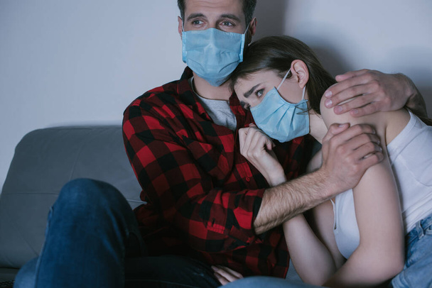 young couple in medical masks watching tv while man hugging sad girl - Foto, imagen