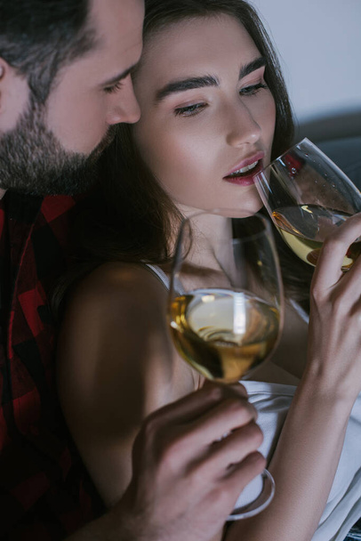young man embracing dreamy girlfriend while sitting on sofa with glasses of white wine - Foto, immagini