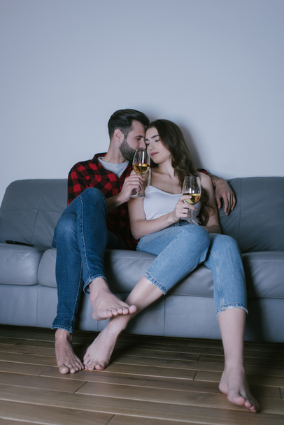 glückliches junges Paar sitzt auf Sofa von Angesicht zu Angesicht und hält Gläser mit Weißwein - Foto, Bild