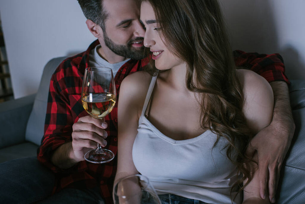 handsome man embracing happy girlfriend while sitting on sofa with glasses of white wine - Fotoğraf, Görsel