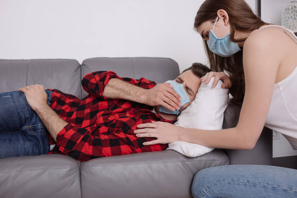 giovane donna in maschera medica accovacciata vicino fidanzato malato sdraiato sul divano in maschera medica e starnuti
 - Foto, immagini