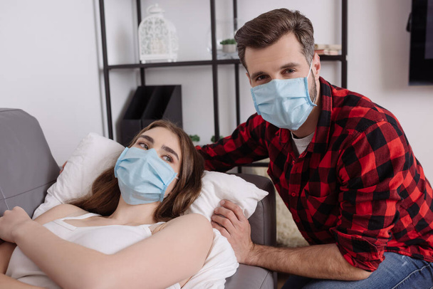 Tıbbi maskeli yakışıklı adam hasta kız arkadaşının yanındaki kanepede uzanan kameraya bakıyor. - Fotoğraf, Görsel