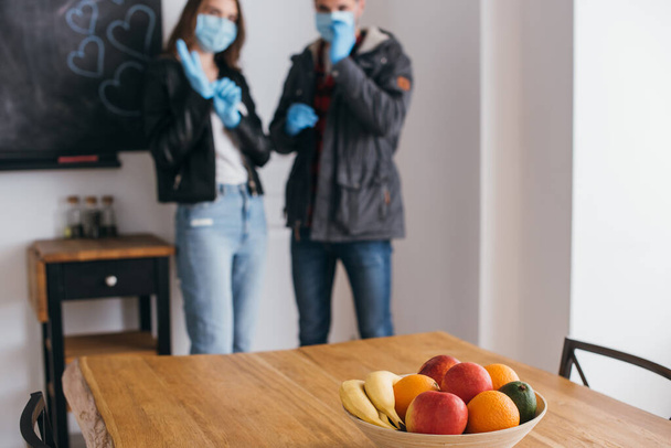 enfoque selectivo de la pareja joven en máscaras médicas y guantes de látex, y tazón de frutas frescas en la mesa
 - Foto, Imagen