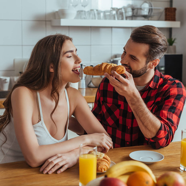 handsome man feeding attractive girlfriend with delicious croissant near orange juice and fresh fruits - Foto, Imagem