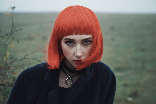 Portrait of beautiful fashion girl with red bob haircut and black clothes in dramatic cold foggy autumn field, young female with the bush with berries and natural landscape on the background - Photo, Image