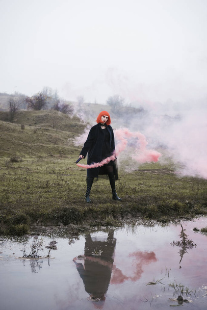Divat lány piros Bob frizura és vörös füst bomba drámai hideg köd őszi területen, fiatal nő vörös füst a természetes táj - Fotó, kép