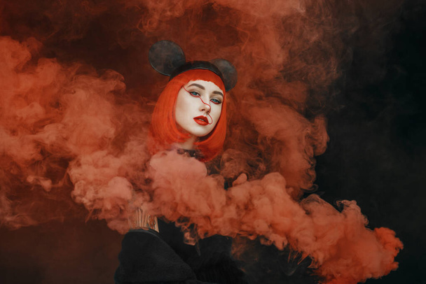 Porträt der hübschen Modeschöpferin mit rotem Bob-Haarschnitt und Mausmütze in dickem roten Rauch, junge Frau mit Mausmütze und Rauchbombe mit dunklem Hintergrund - Foto, Bild