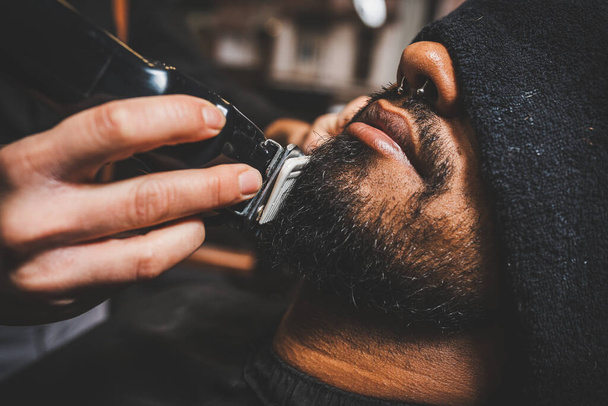 un barbero esta pasando la maquina elctrica de cortar y afeitar por la barba de un chico brasileo afroamericano. ντε εστίλο σουίνγκ χίπστερ. Estn en una Barbera, πελουκέρα. - Φωτογραφία, εικόνα