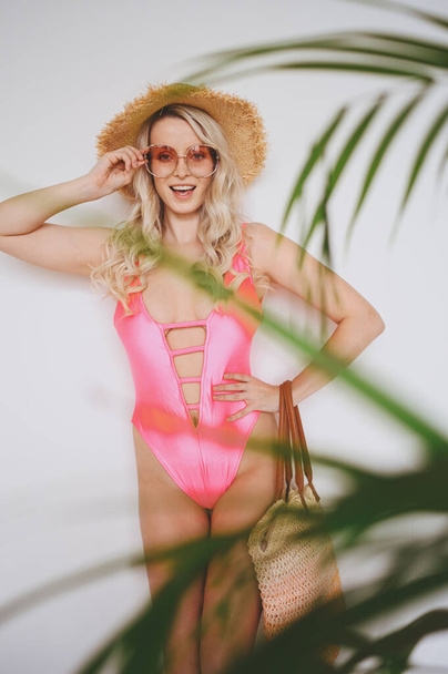 Young sexy smiling happy blonde in a pink swimsuit, a straw hat, sunglasses, excited to present product. Woman on a white background with palm green leaves. Summer sales concept copy space mock up - Foto, Imagem