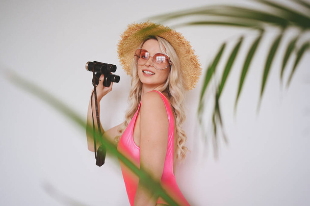 Junge sexy lächelnde glückliche Blondine in rosa Badeanzug, Strohhut, Sonnenbrille, aufgeregt, Produkt zu präsentieren. Frau auf weißem Hintergrund mit palmengrünen Blättern. Sommerschlussverkauf-Konzept kopiert Fläche - Foto, Bild