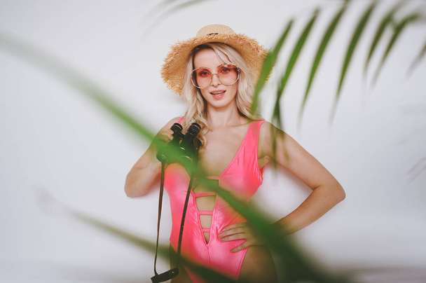 Jovem sexy sorrindo loira feliz em um maiô rosa, um chapéu de palha, óculos de sol, animado para apresentar o produto. Mulher sobre um fundo branco com folhas de palma verde. Verão vendas conceito cópia espaço simular
 - Foto, Imagem
