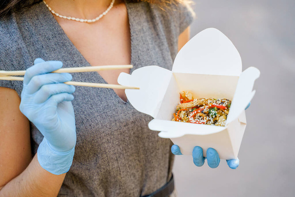 Primo piano cibo gustoso wok nella consegna scatola. Ragazza tiene tagliatelle udon con tempuru, gamberetti, salsa di soia in mano in guanti medici. Cibo piccante giapponese in scatole eco usa e getta. Wok scatola udon pubblicizzare
 - Foto, immagini
