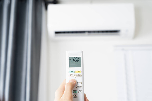 A mão humana está usando o controle remoto branco do condicionador de ar para ligar ou ajustar a temperatura do condicionador de ar dentro da sala. - Foto, Imagem