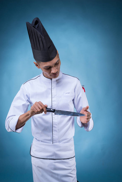 Un retrato de un chef ejecutivo asiático con su uniforme blanco y delantal, sin su sombrero sobre fondo azul jugando con su cuchillo
 - Foto, Imagen