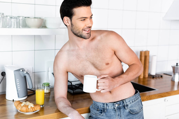 veselý muž bez košile drží šálek kávy na kuchyni se snídaní - Fotografie, Obrázek