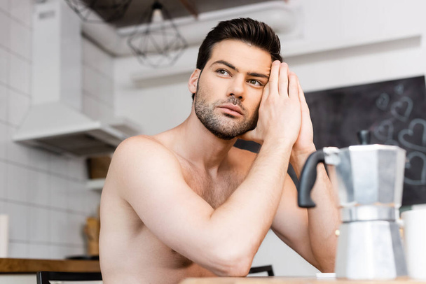 doordachte shirtloze trieste man zittend op keuken met koffiepot - Foto, afbeelding