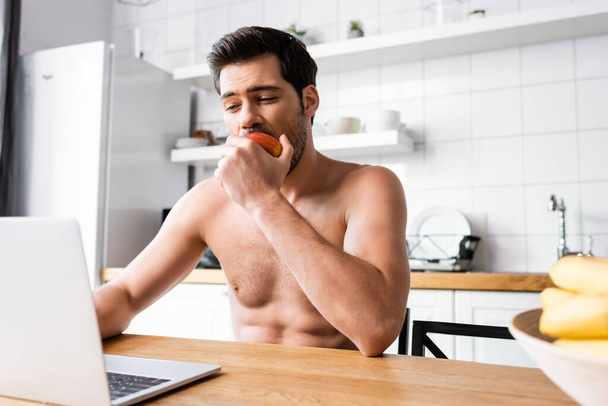 handsome shirtless freelancer eating apple while working on laptop on kitchen - Fotoğraf, Görsel