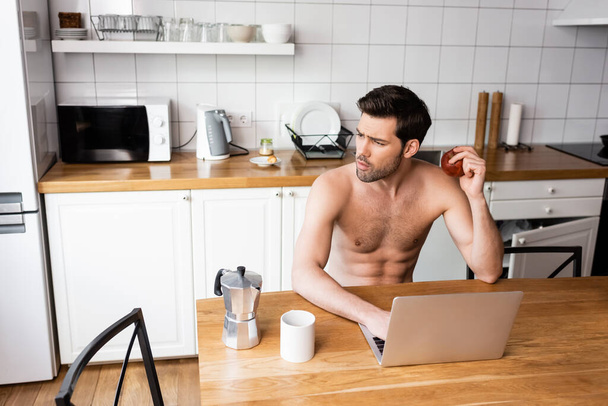 thoughtful freelancer eating apple while working on laptop on kitchen - Foto, Imagem