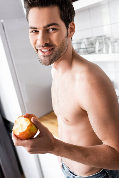 happy shirtless man eating apple on kitchen in morning - Foto, Imagen