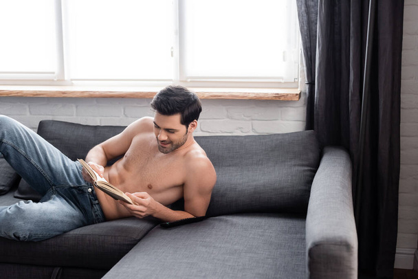 pohledný usměvavý muž bez košile čtení knihy na pohovce - Fotografie, Obrázek