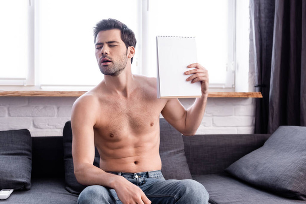 сексуальный мужчина без рубашки с закрытыми глазами страдает от жары, размахивая блокнотом в качестве вентилятора дома
 - Фото, изображение