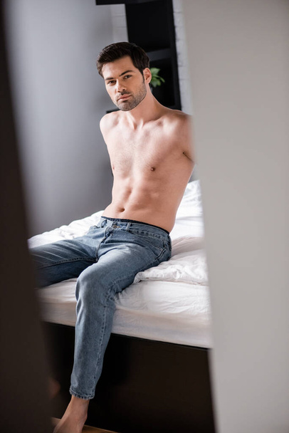 schöner sexy hemdloser Mann in Jeans sitzt auf dem Bett, selektiver Fokus - Foto, Bild