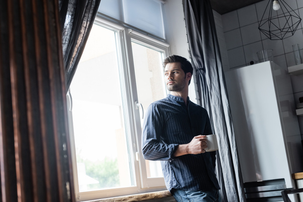 bonito homem pensativo segurando xícara de café e de pé perto da janela em casa em auto-isolamento
 - Foto, Imagem