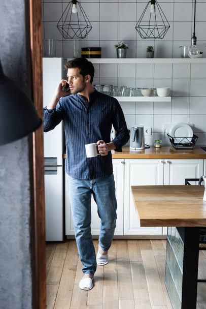 nachdenklicher Mann mit Tasse Kaffee, während er während der Quarantäne mit dem Handy in der Küche spricht   - Foto, Bild