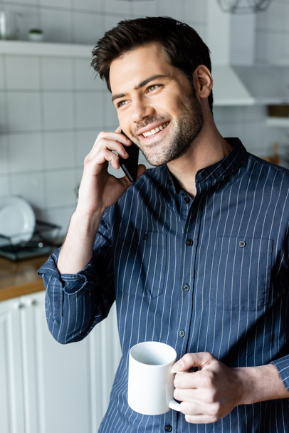 Karantina sırasında mutfakta telefonla konuşurken elinde kahve tutan neşeli yakışıklı bir adam.   - Fotoğraf, Görsel