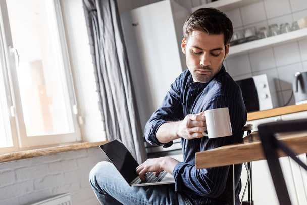 handsome man holding cup of coffee while using laptop on kitchen during quarantine   - Φωτογραφία, εικόνα