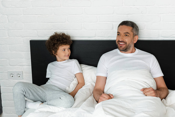 curly boy looking at bearded father in bedroom  - Фото, изображение