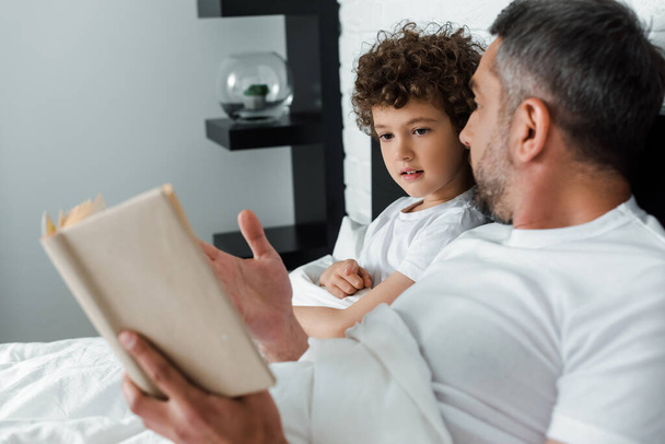 selective focus of curly boy looking at book near father in bedroom  - 写真・画像