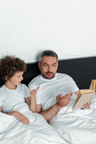 bearded father reading book near cute son in bedroom  - Zdjęcie, obraz
