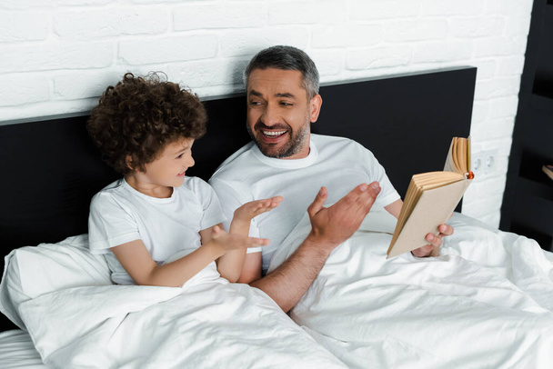 curly boy gesturing near happy father holding book in bedroom  - Fotoğraf, Görsel