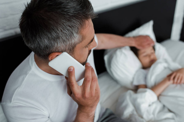 selective focus of father touching sick son and talking on smartphone in bedroom  - Photo, Image
