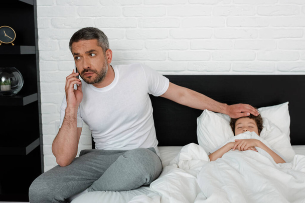 γενειοφόρος πατέρας αγγίζοντας μέτωπο του άρρωστου γιου και μιλώντας στο smartphone - Φωτογραφία, εικόνα